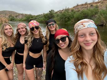Six friends wearing colorful bandanas and black activewear take a smiling selfie on a rocky riverbank with green brush and arid hills in the background, outdoor river trip