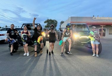 Group of six adults in helmets and casual gear posing at dusk in a small-town parking lot beside two off-road UTVs with headlights on, ready for an evening trail ride.