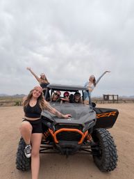 Six friends posing on an orange-accented off-road UTV on a dusty desert trail, smiling and raising their arms beneath a cloudy sky with distant mountains — outdoor off-road adventure.