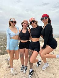 Four smiling women in bandanas and athletic outfits pose on a rocky desert hilltop overlook above a suburban valley — playful hiking group photo.