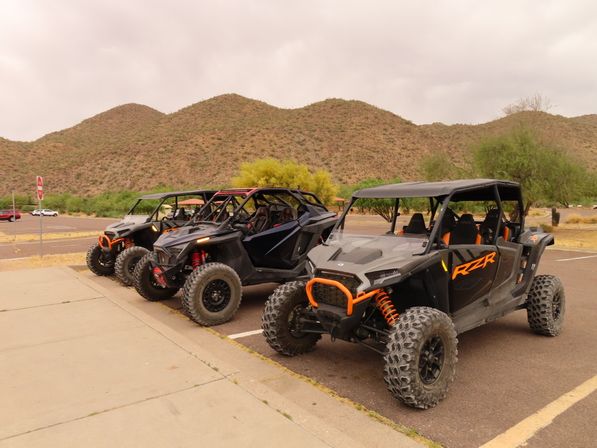 Three rugged off-road UTV side-by-sides with chunky tires and orange accents parked in a desert lot against scrubby foothills and a cloudy sky.