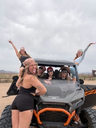 Group of friends posing and making faces on and around an orange-accented off-road UTV on a dusty desert trail with cloudy sky and distant mountains — outdoor off-road adventure in the Southwest.