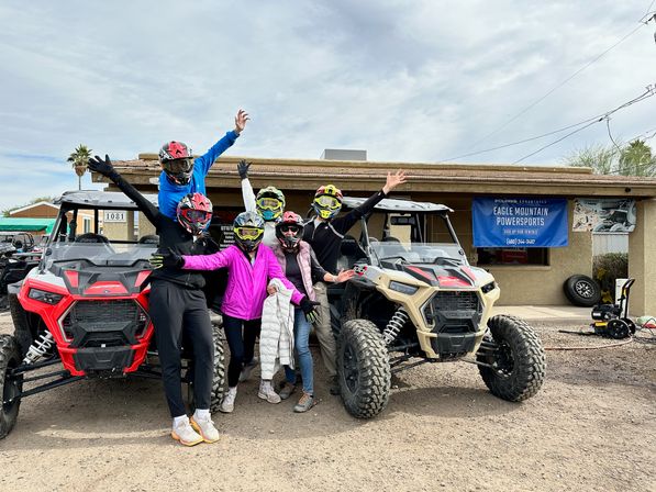 Six helmeted off-road riders in colorful jackets posing with arms raised between red and tan UTV side-by-sides parked in front of a desert storefront under a cloudy sky.