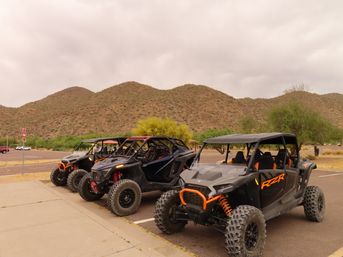 Three orange-accented off-road UTVs parked at a desert trailhead lot under an overcast sky, with scrubby vegetation and rounded desert hills in the background.