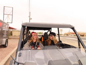 Two women wearing bandanas, sunglasses and safety harnesses seated in a side-by-side off-road UTV parked near a roadside shopping strip in a desert town