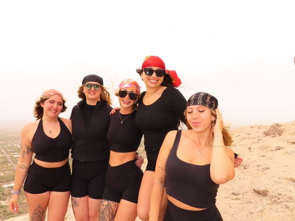Five friends in black activewear and colorful bandanas smiling together on a rocky desert hilltop, sunglasses and tattoos visible, with a hazy valley view below — outdoor hiking group.
