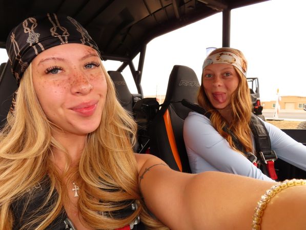 Playful selfie of two women in an off-road side-by-side vehicle wearing bandanas and harnesses, one smiling and the other sticking out her tongue against a desert backdrop.