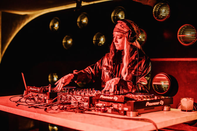 DJ mixing a live set at a red-lit nightclub: person in a bandana and headphones adjusting a multi-deck mixer on a table with glowing circular stage lights and a cocktail glass nearby.