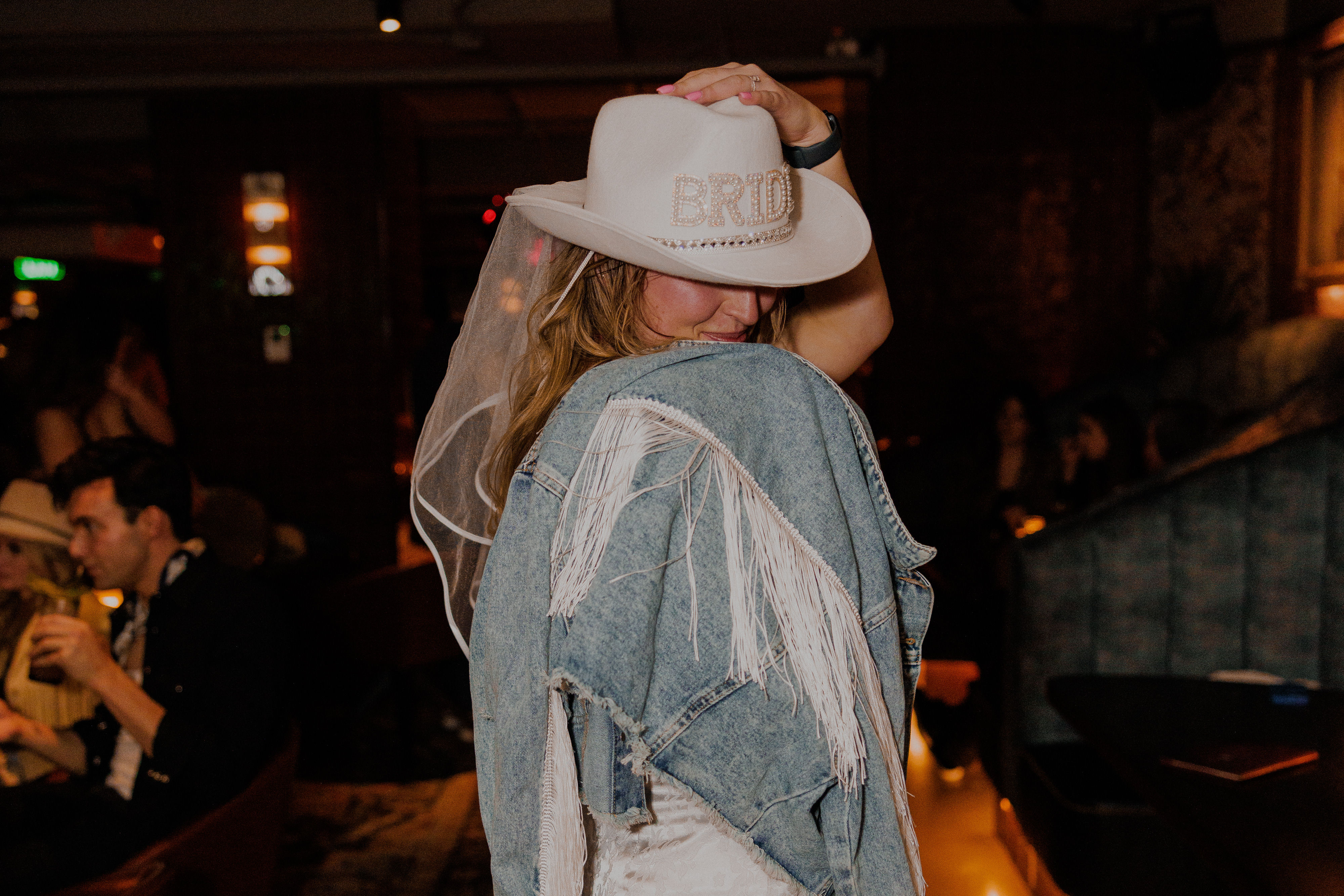 Bride wearing a white 'BRIDE' cowboy hat and veil, wrapped in a fringed denim jacket, posing in a dimly lit bar lounge at a bachelorette party.