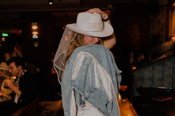 Bride wearing a white 'BRIDE' cowboy hat and veil, wrapped in a fringed denim jacket, posing in a dimly lit bar lounge at a bachelorette party.