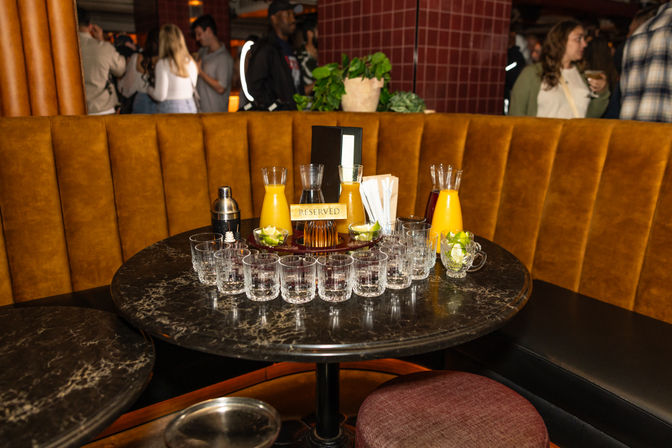 Reserved marble cocktail table at a busy lounge bar with mustard velvet booth, tray of empty crystal glasses arranged in a semicircle, carafes of orange juice, lime wedges and a cocktail shaker