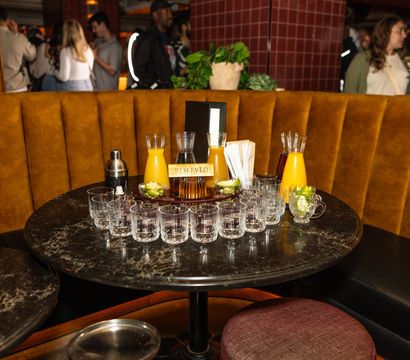 Reserved marble cocktail table at a busy lounge bar with mustard velvet booth, tray of empty crystal glasses arranged in a semicircle, carafes of orange juice, lime wedges and a cocktail shaker