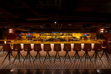 Moody underground cocktail bar interior with a long backlit amber counter, a row of brown leather swivel stools, patterned tile floor and illuminated liquor shelves — warm urban speakeasy vibe.