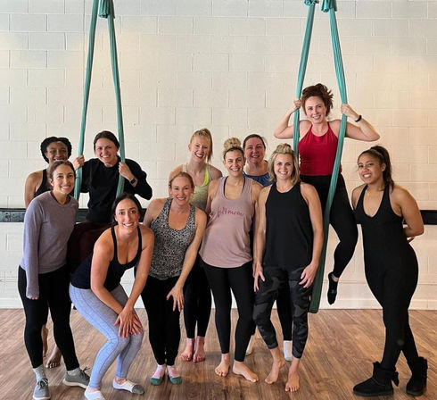 Smiling group of women in activewear posing with teal aerial silks in an indoor fitness studio — aerial yoga class on hardwood floor with white brick wall