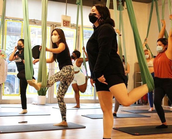 Group of masked adults balancing and stretching in an aerial yoga class using green hammocks and mats in a bright fitness studio.