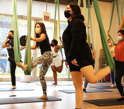 Group of masked adults balancing and stretching in an aerial yoga class using green hammocks and mats in a bright fitness studio.