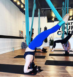 Aerial yoga class in an indoor studio with turquoise silks — participant in blue leggings suspended upside down in an inversion over a black mat on a hardwood floor.