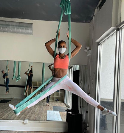 Person wearing a face mask, coral sports bra and white leggings performing a wide-leg split suspended in teal aerial silk inside an indoor aerial yoga studio with mirrors and large windows.