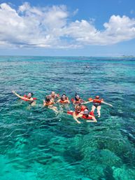 Group of snorkelers in orange life vests floating above clear turquoise coral reef water in a tropical ocean under a sunny blue sky with fluffy clouds