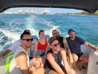 Smiling group of friends on a motorboat cruising over turquoise tropical water, boat wake trailing behind and large docked cruise ships on the horizon under a sunny sky.
