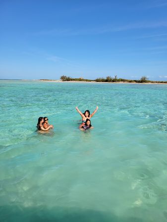 Four people splashing and posing in shallow turquoise ocean near a sandy tropical island under a clear blue sky, sunny beach scene