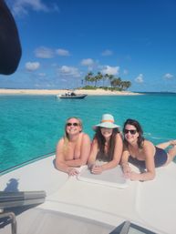 Three smiling women in swimsuits lounging on the bow of a white boat in crystal turquoise water near a palm‑fringed sandbar with a small motorboat and clear blue sky.