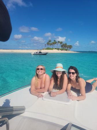 Three smiling women in swimsuits lounging on the bow of a white boat in crystal turquoise water near a palm‑fringed sandbar with a small motorboat and clear blue sky.