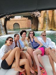Five friends laughing on a small boat during a summer boat tour in a resort-style waterway, sitting on a cushioned bench with a watermelon inflatable underfoot and ornate stone arch with cascading waterfalls in the background.