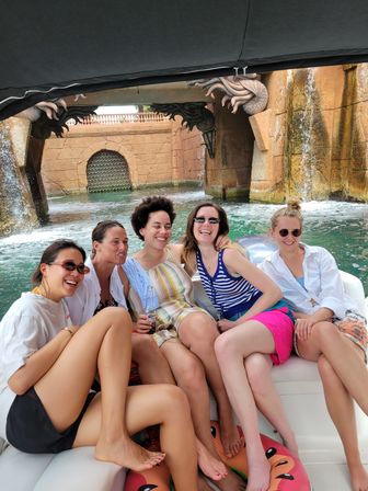 Five friends laughing on a small boat during a summer boat tour in a resort-style waterway, sitting on a cushioned bench with a watermelon inflatable underfoot and ornate stone arch with cascading waterfalls in the background.