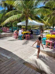 Sunny tropical beachside deck with palm trees, colorful picnic tables and stools, an umbrella-shaded group dining area, and kids playing on the wooden boardwalk.