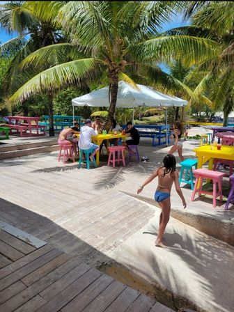 Sunny tropical beachside deck with palm trees, colorful picnic tables and stools, an umbrella-shaded group dining area, and kids playing on the wooden boardwalk.