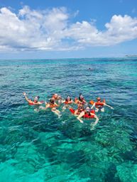 Group of snorkelers in orange life vests floating over a turquoise coral reef in crystal-clear tropical ocean under a sunny blue sky with puffy clouds