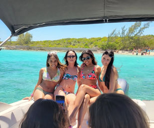 Four friends in colorful bikinis posing on a boat with turquoise tropical water and a sandy shoreline in the background as someone snaps a photo.