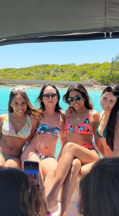 Four friends in colorful bikinis posing on a boat with turquoise tropical water and a sandy shoreline in the background as someone snaps a photo.