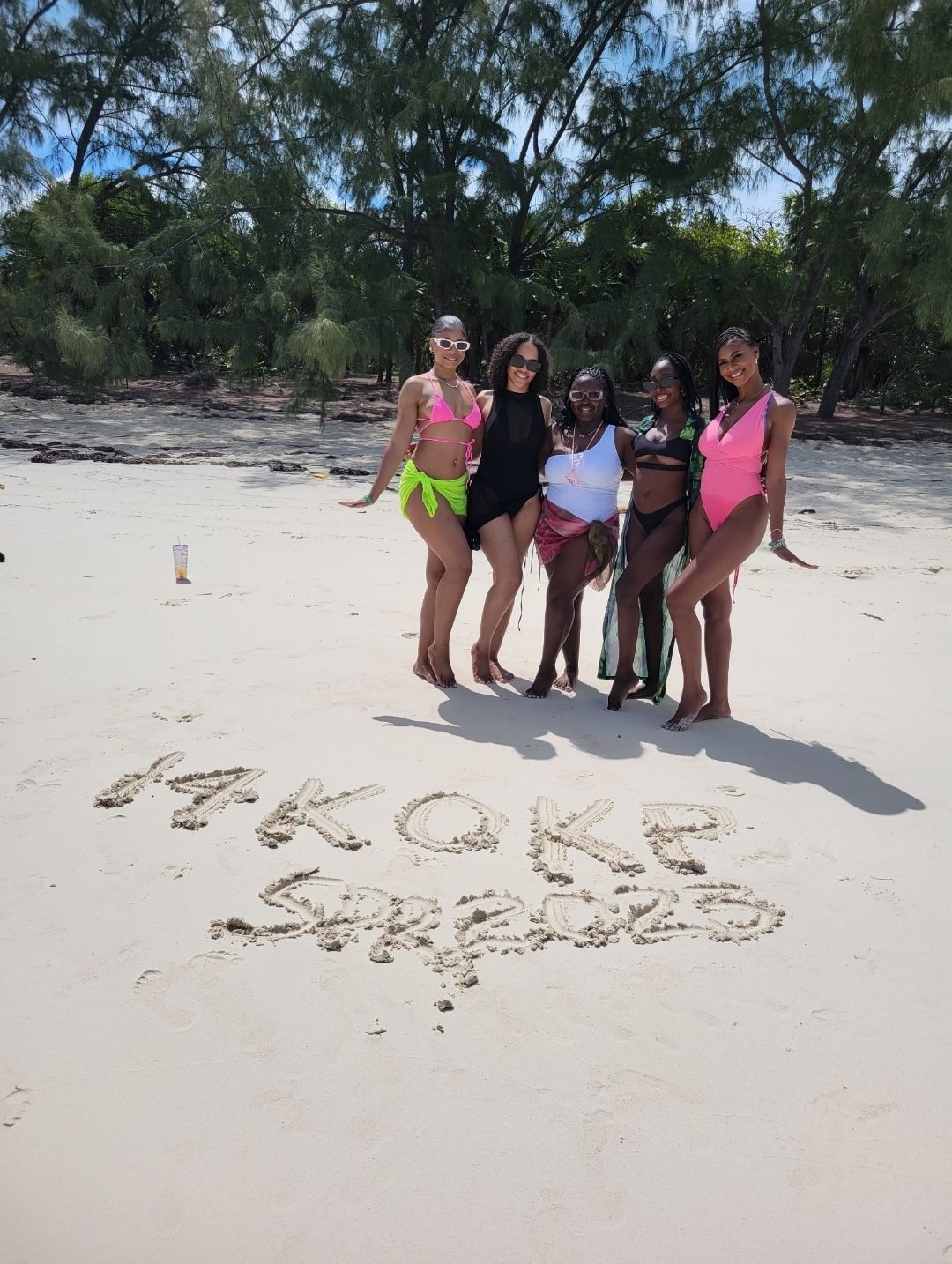 Five women in colorful swimsuits posing on a sunny white-sand tropical beach with a tree-lined shore and words written in the sand, vacation group photo.