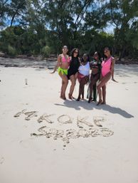 Five women in colorful swimsuits posing on a sunny white-sand tropical beach with a tree-lined shore and words written in the sand, vacation group photo.
