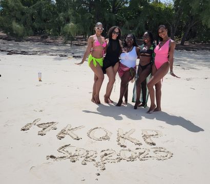 Five women in colorful swimsuits posing on a sunny white-sand tropical beach with a tree-lined shore and words written in the sand, vacation group photo.