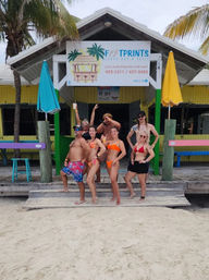 Seven people in swimsuits posing on the wooden steps of a colorful tropical beach bar with palm trees, bright umbrellas and sandy shoreline.
