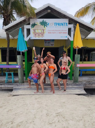 Seven people in swimsuits posing on the wooden steps of a colorful tropical beach bar with palm trees, bright umbrellas and sandy shoreline.