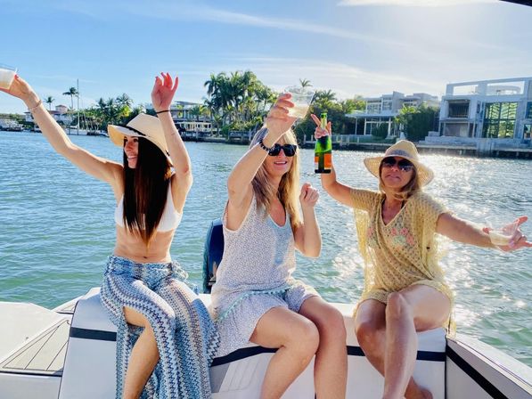 Three women celebrating on a boat, wearing sun hats and sunglasses, toasting with champagne and drinks by a palm-lined waterfront under a sunny sky