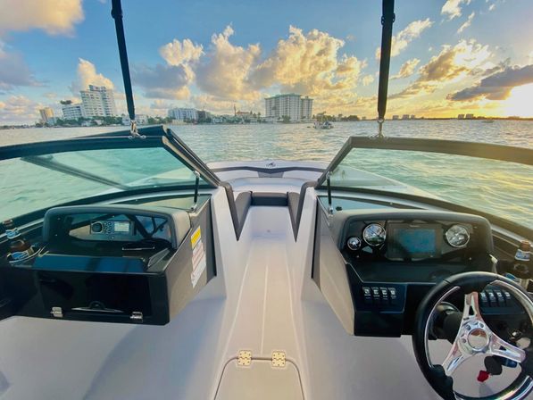View from a motorboat cockpit over the bow with steering wheel and gauges, turquoise bay and waterfront city skyline glowing under a golden sunset.