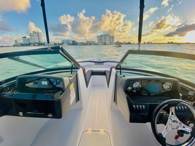 View from a motorboat cockpit over the bow with steering wheel and gauges, turquoise bay and waterfront city skyline glowing under a golden sunset.