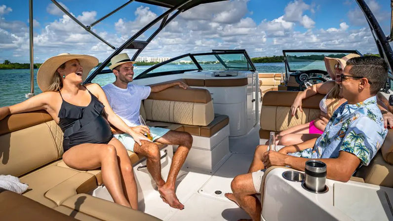 Four friends laughing on a sunny motorboat cruise over turquoise coastal waters, relaxing on tan leather seats with summer hats and drinks under a blue sky with fluffy clouds.