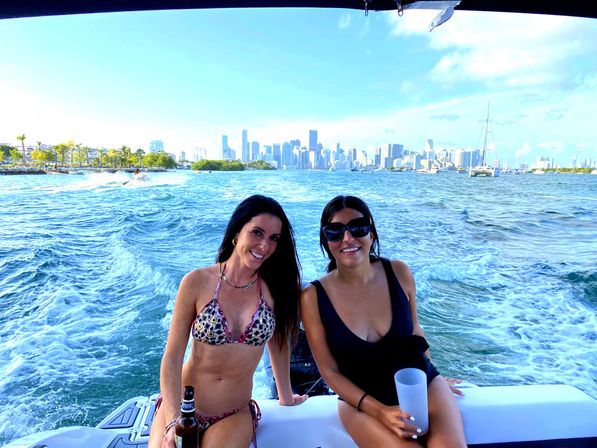 Two friends in swimsuits smiling aboard a boat in Miami’s Biscayne Bay, city skyline and palm-lined shore behind them with sailboats and wake in turquoise water.