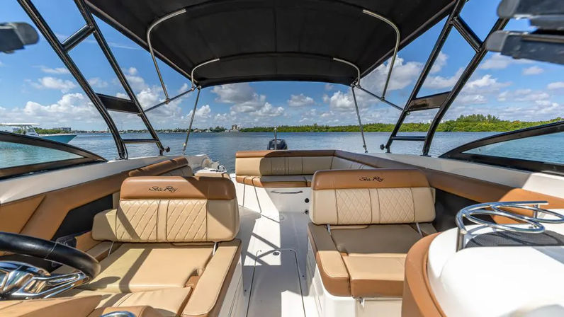 Spacious tan-leather motorboat cockpit with cushioned seats under a black bimini, looking out over calm blue water and a tree-lined shoreline under a sunny sky