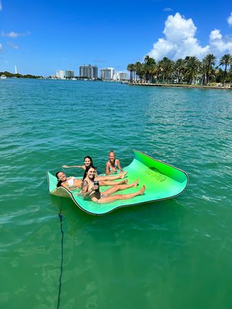 Four people lounging on a bright green floating mat in clear turquoise bay, sipping drinks with a palm-lined shoreline and high-rise city skyline under a sunny blue sky.