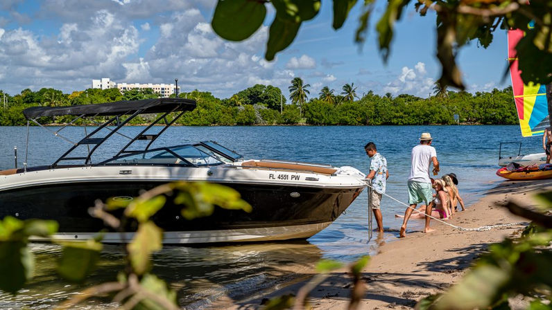 People securing a black-and-white motorboat on a sandy Florida inlet while kids play at the water’s edge, with palm trees and a colorful sailboat in the background under sunny skies.