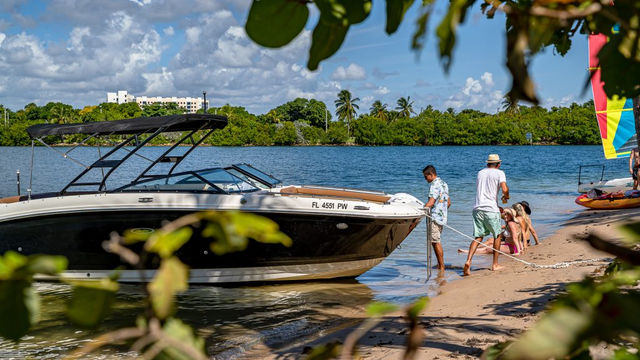 People securing a black-and-white motorboat on a sandy Florida inlet while kids play at the water’s edge, with palm trees and a colorful sailboat in the background under sunny skies.