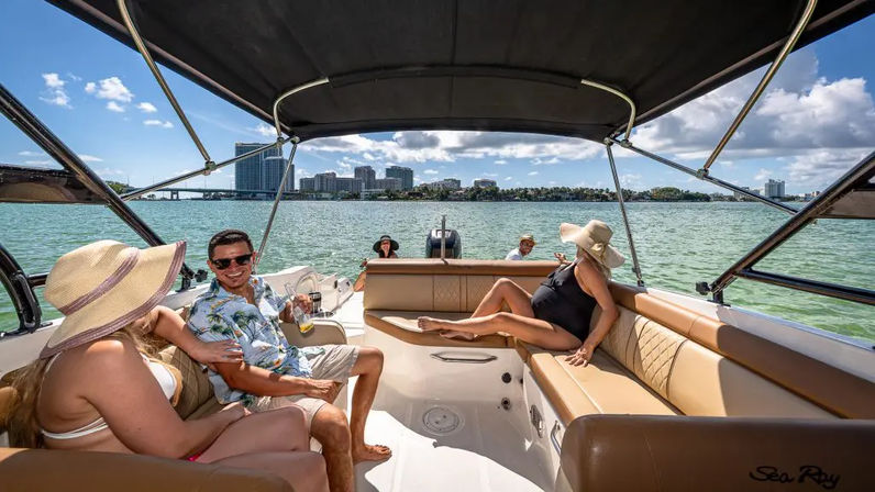 Smiling friends lounging on a sunny motorboat under a canopy, sipping drinks and enjoying a coastal city skyline across a turquoise bay.