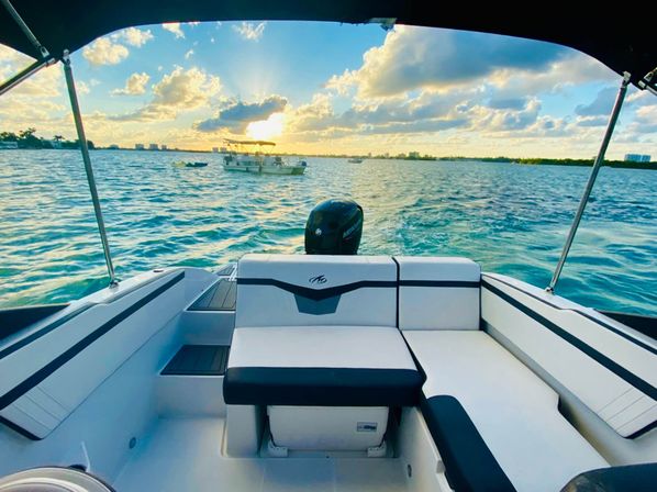 Sunset view from a motorboat's stern seating over turquoise coastal bay, sun-kissed waves, small boats and city skyline on the horizon.